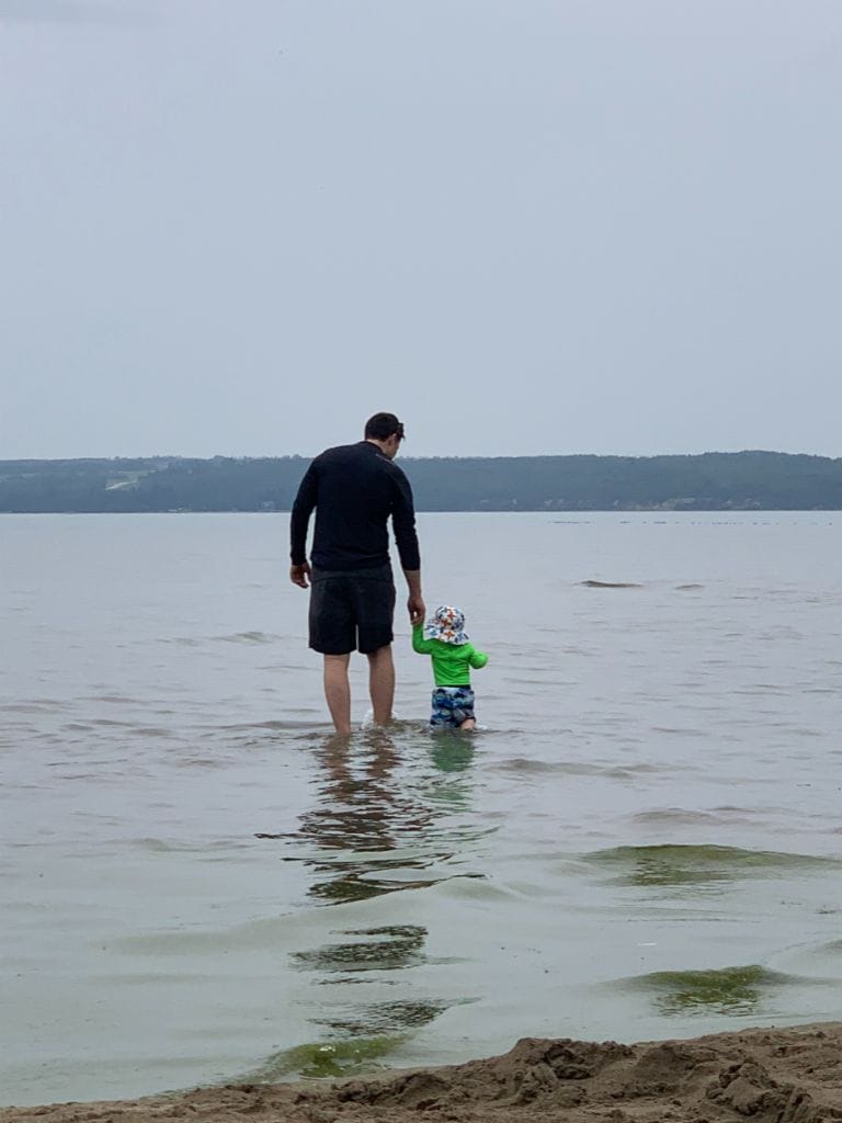 A father and his child walking into a lake with a beach.